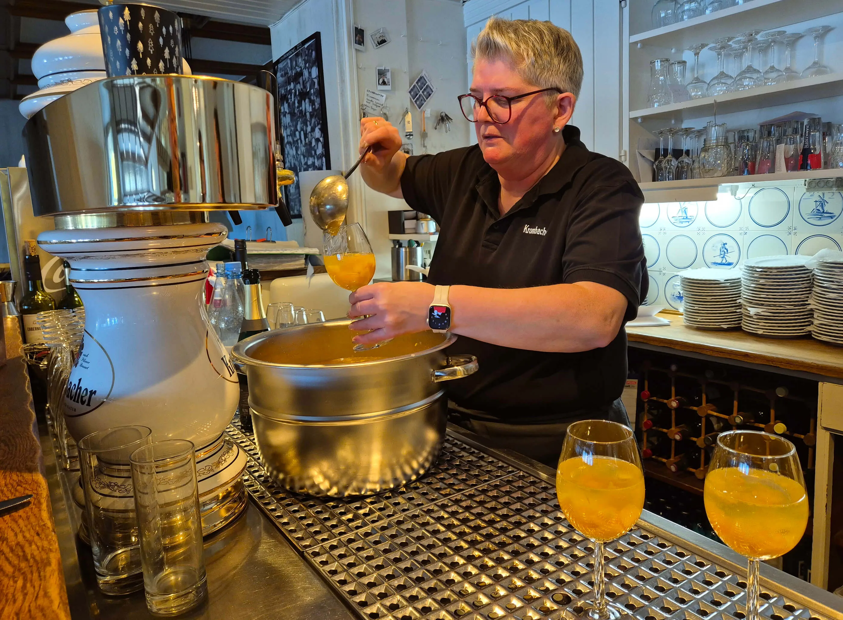 Ein Mann mit kurzem grauen Haar schöpft mit einer Suppenkelle eine orangefarbene Flüssigkeit aus einem großen Topf in ein Glas. Im Hintergrund steht eine große metallene Brennerei und Regale mit Gläsern.