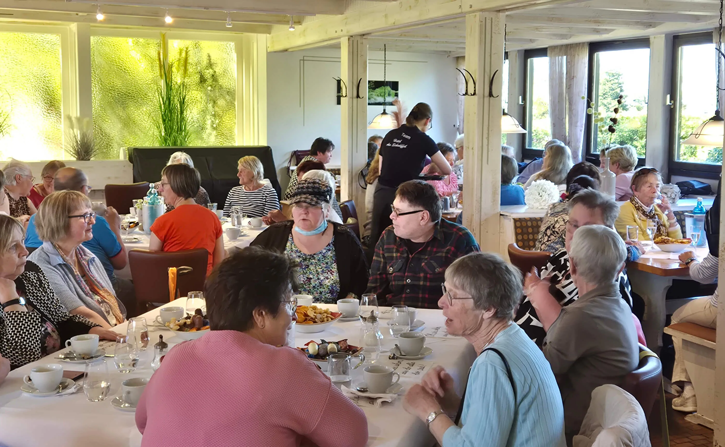 Mehrere Tische in einem hellen Restaurant sind mit Menschen unterschiedlichen Alters gefüllt, die sich unterhalten und Speisen von Tellern essen. Im Hintergrund bedient ein Mitarbeiter.