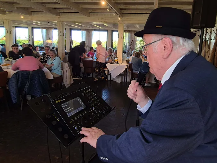 Ein älterer Mann mit Hut und Hemd spricht in ein Mikrofon, während er an einem Mischpult mit Touchscreen-Display steht. Im Hintergrund sitzen Menschen an Tischen in einem hellen Restaurant.