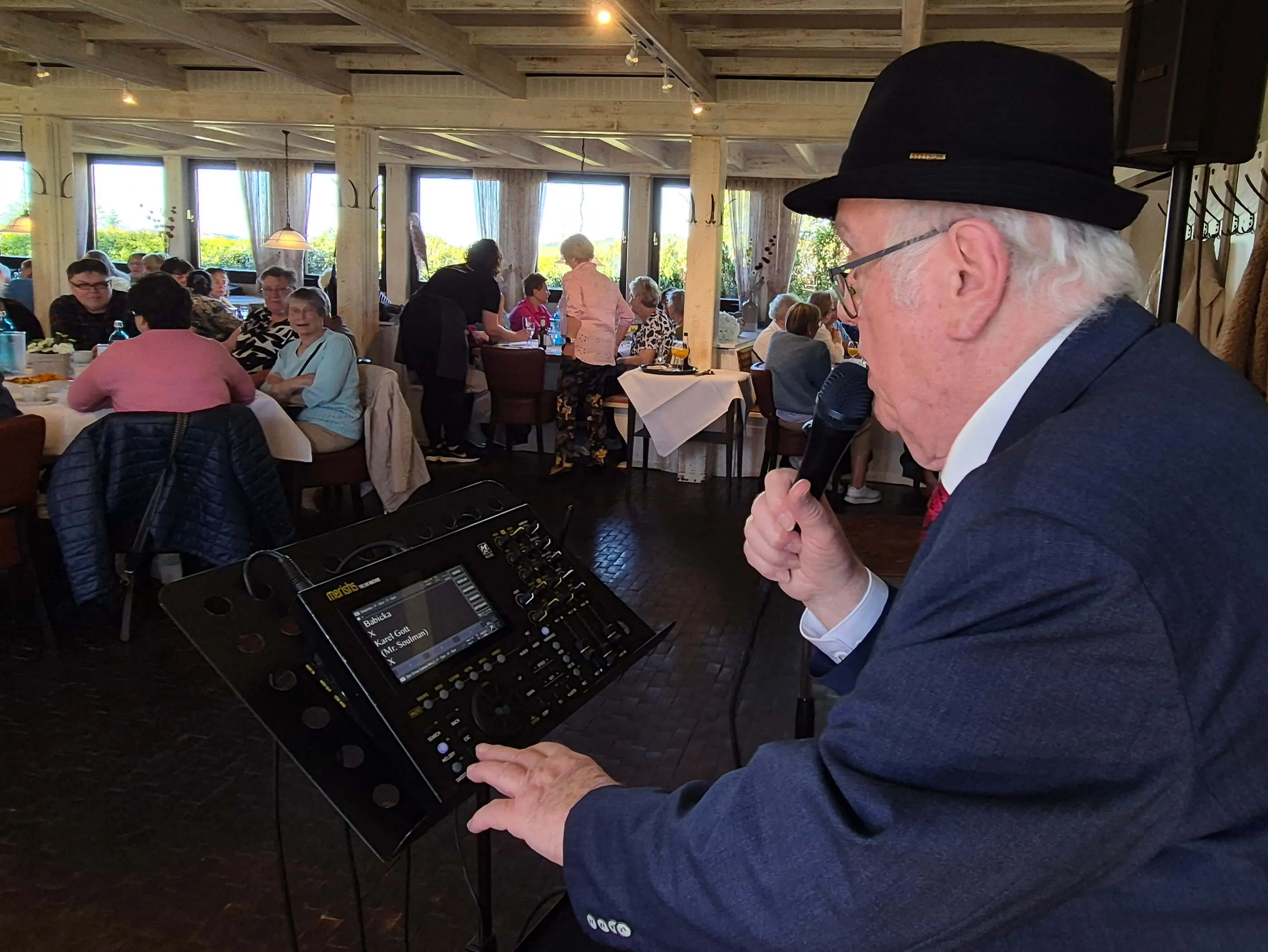 Ein älterer Mann mit Hut und Hemd spricht in ein Mikrofon, während er an einem Mischpult mit Touchscreen-Display steht. Im Hintergrund sitzen Menschen an Tischen in einem hellen Restaurant.