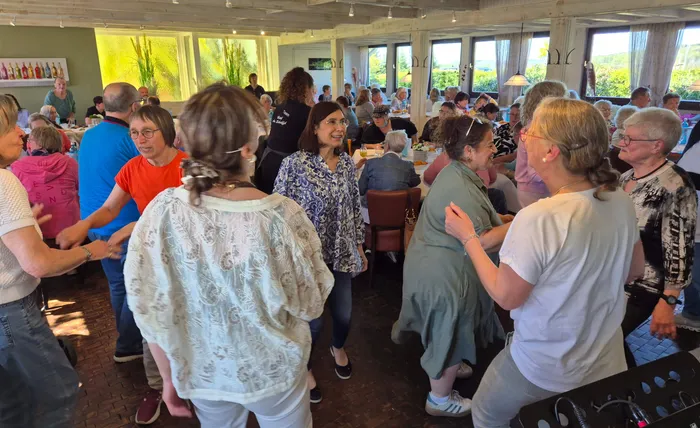 Eine Gruppe älterer Menschen tanzt im hellen Restaurant, während andere Gäste an Tischen sitzen und zusehen. Im Hintergrund sind große Fenster mit Blick auf die Landschaft zu sehen.