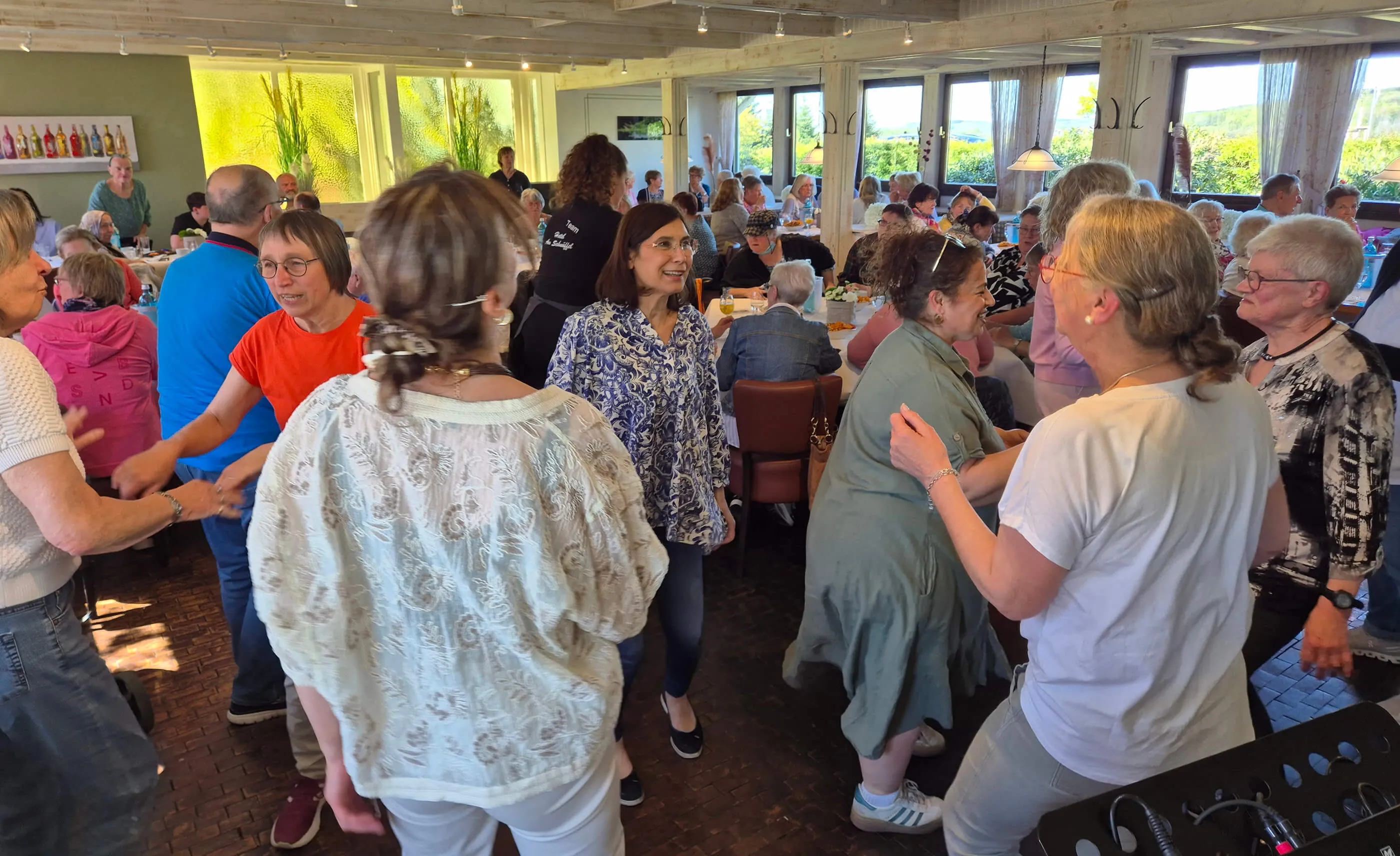Eine Gruppe älterer Menschen tanzt im hellen Restaurant, während andere Gäste an Tischen sitzen und zusehen. Im Hintergrund sind große Fenster mit Blick auf die Landschaft zu sehen.