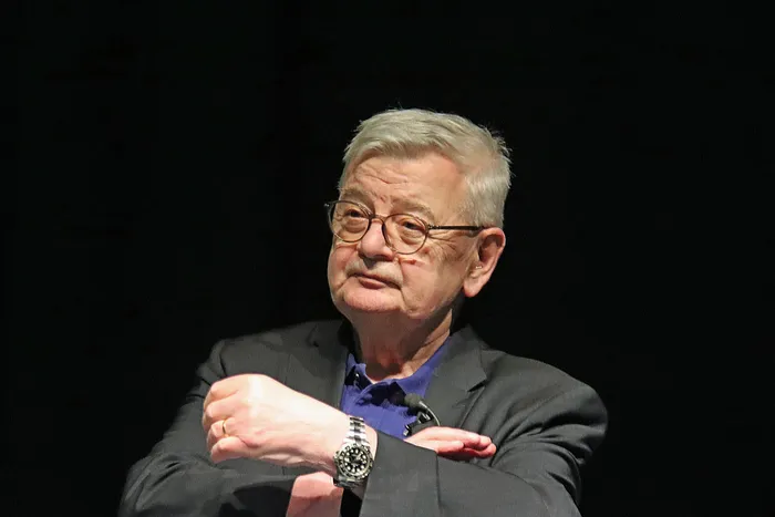 Joschka Fischer, ein älterer Mann mit weißem Haar und runder Brille, blickt nachdenklich zur Seite. Er trägt Anzug und blaues Hemd und hat die Arme vor dem Körper verschränkt.