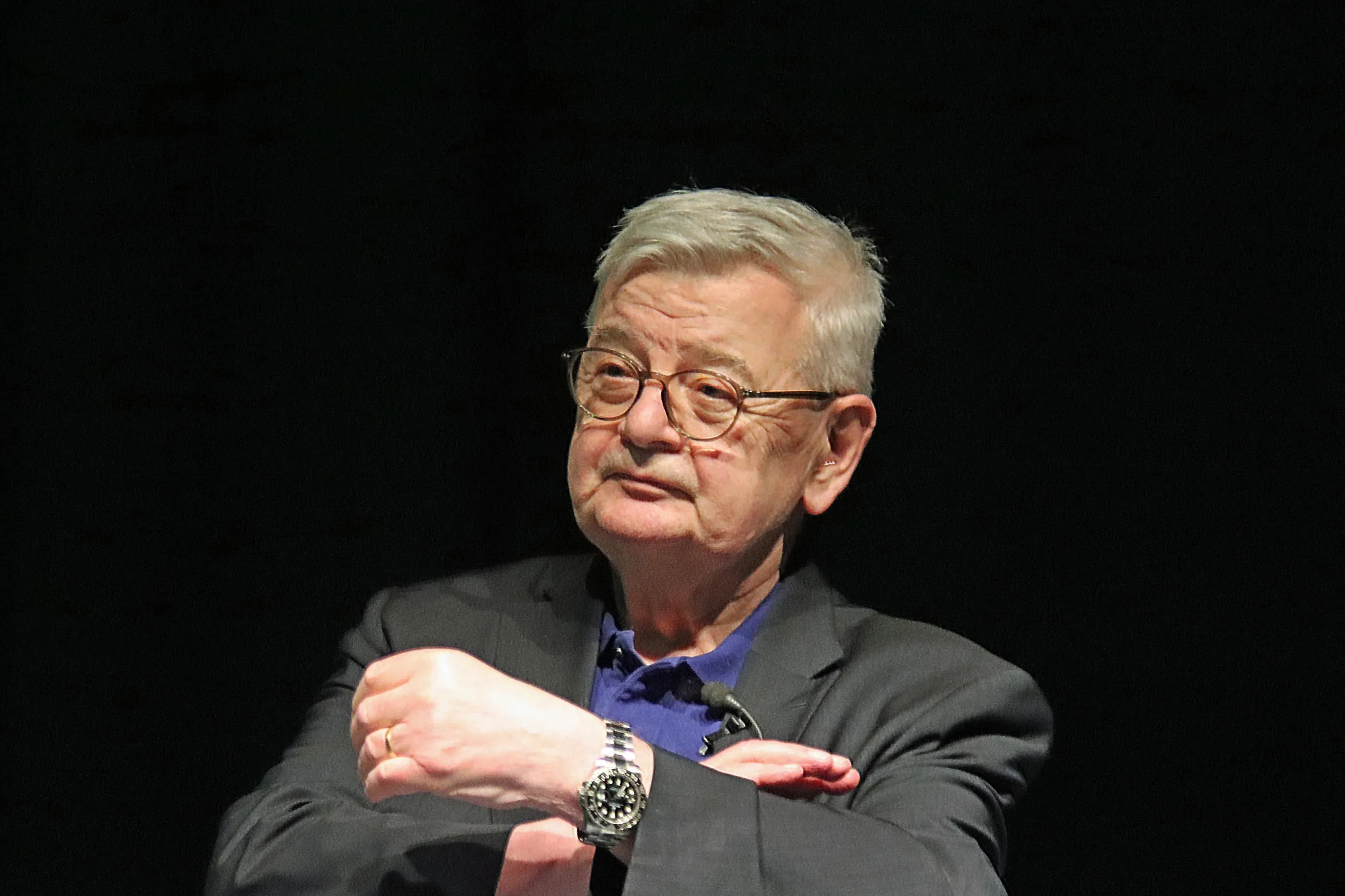 Joschka Fischer, ein älterer Mann mit weißem Haar und runder Brille, blickt nachdenklich zur Seite. Er trägt Anzug und blaues Hemd und hat die Arme vor dem Körper verschränkt.