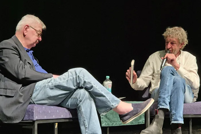 Joschka Fischer und Matthias Bongard sitzen auf Stühlen auf einer Bühne und unterhalten sich. Fischer hält ein Buch in der Hand, Bongard ein Mikrofon.