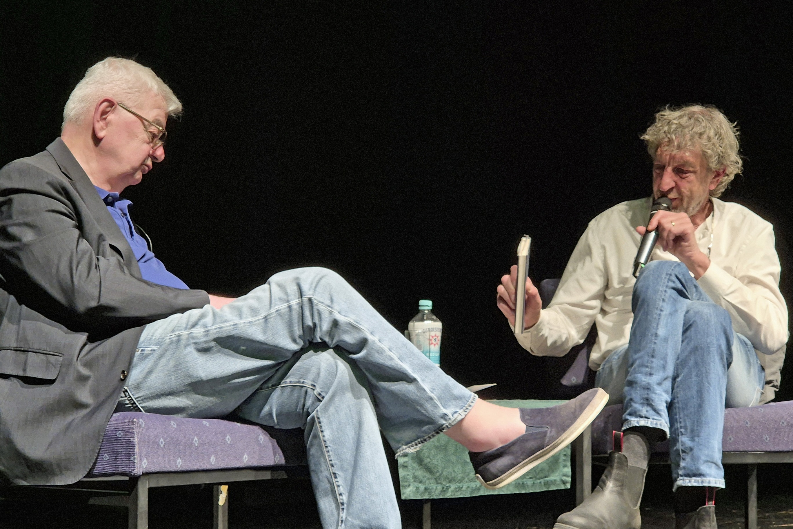 Joschka Fischer und Matthias Bongard sitzen auf Stühlen auf einer Bühne und unterhalten sich. Fischer hält ein Buch in der Hand, Bongard ein Mikrofon.