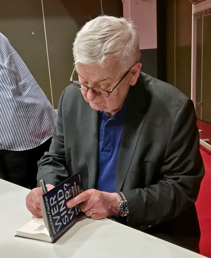 Joschka Fischer, ein älterer Mann mit grauem Haar und Brille, signiert ein Buch mit dem Titel „Wer sind wir?“.