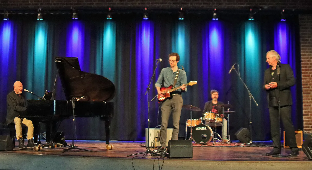 Vier Musiker auf einer Bühne: Ein Mann spielt Klavier, ein anderer E-Gitarre, ein dritter sitzt am Schlagzeug und ein vierter hält ein Mikrofon. Im Hintergrund ein blauer Vorhang und Bühnenlichter.