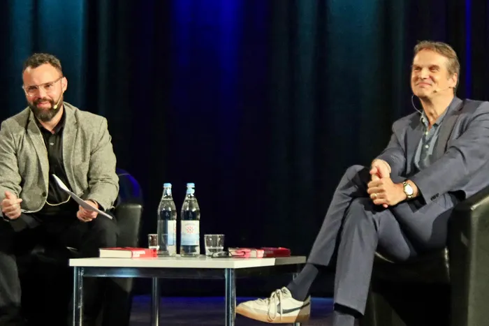 Zwei Männer sitzen an einem Tisch auf einer Bühne und diskutieren. Links trägt einer einen Tweed-Anzug, der andere rechts einen blauen Anzug und Turnschuhe.