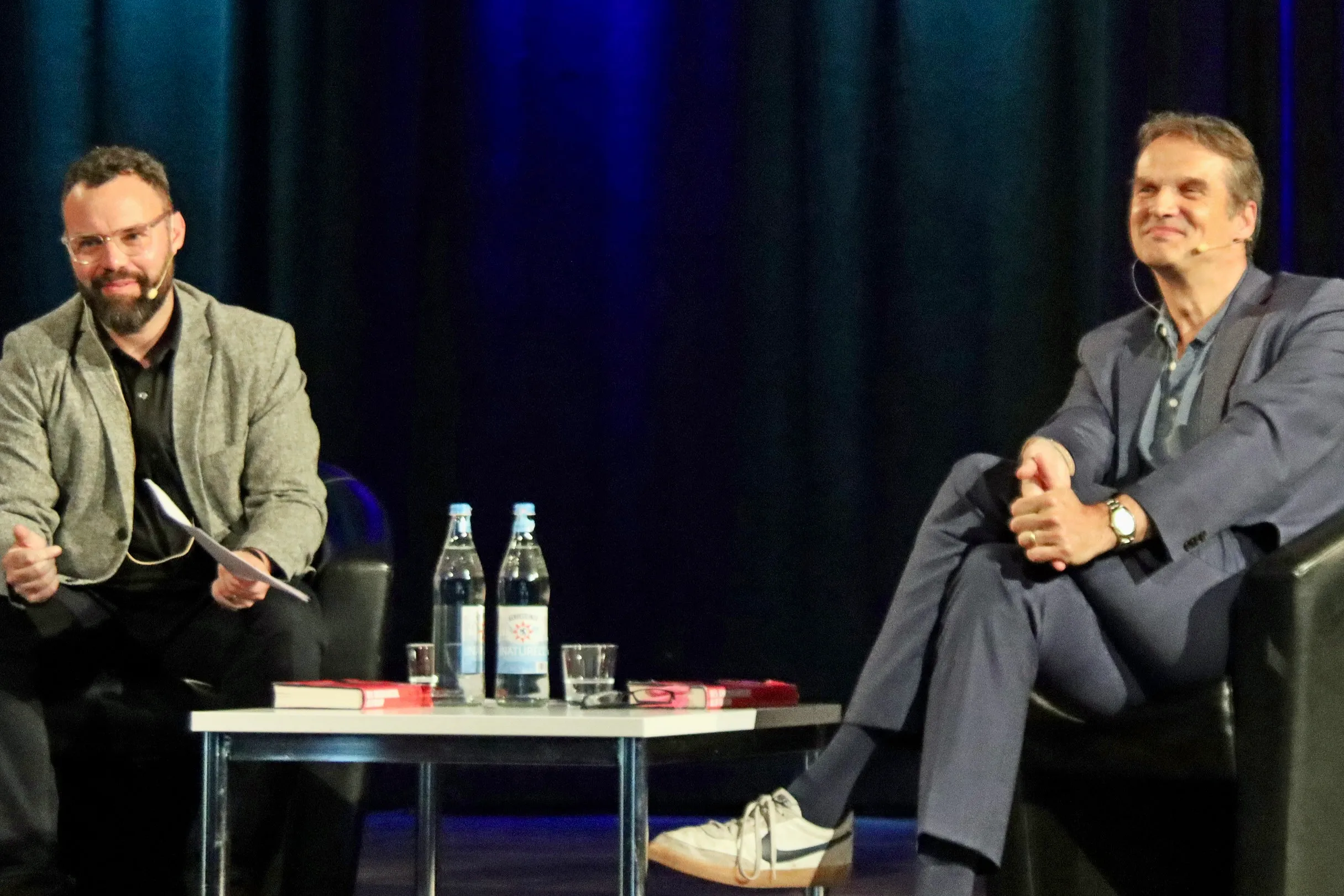 Zwei Männer sitzen an einem Tisch auf einer Bühne und diskutieren. Links trägt einer einen Tweed-Anzug, der andere rechts einen blauen Anzug und Turnschuhe.