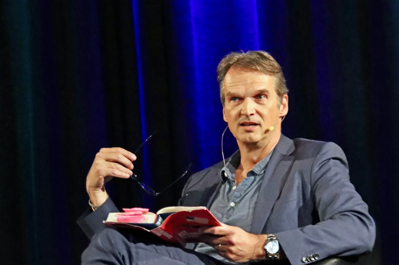 Klaus Brinkbäumer hält während einer Lesung ein Buch in der Hand und blickt ins Publikum. Er trägt Anzug und Hemd und hat eine Brille in der Hand.