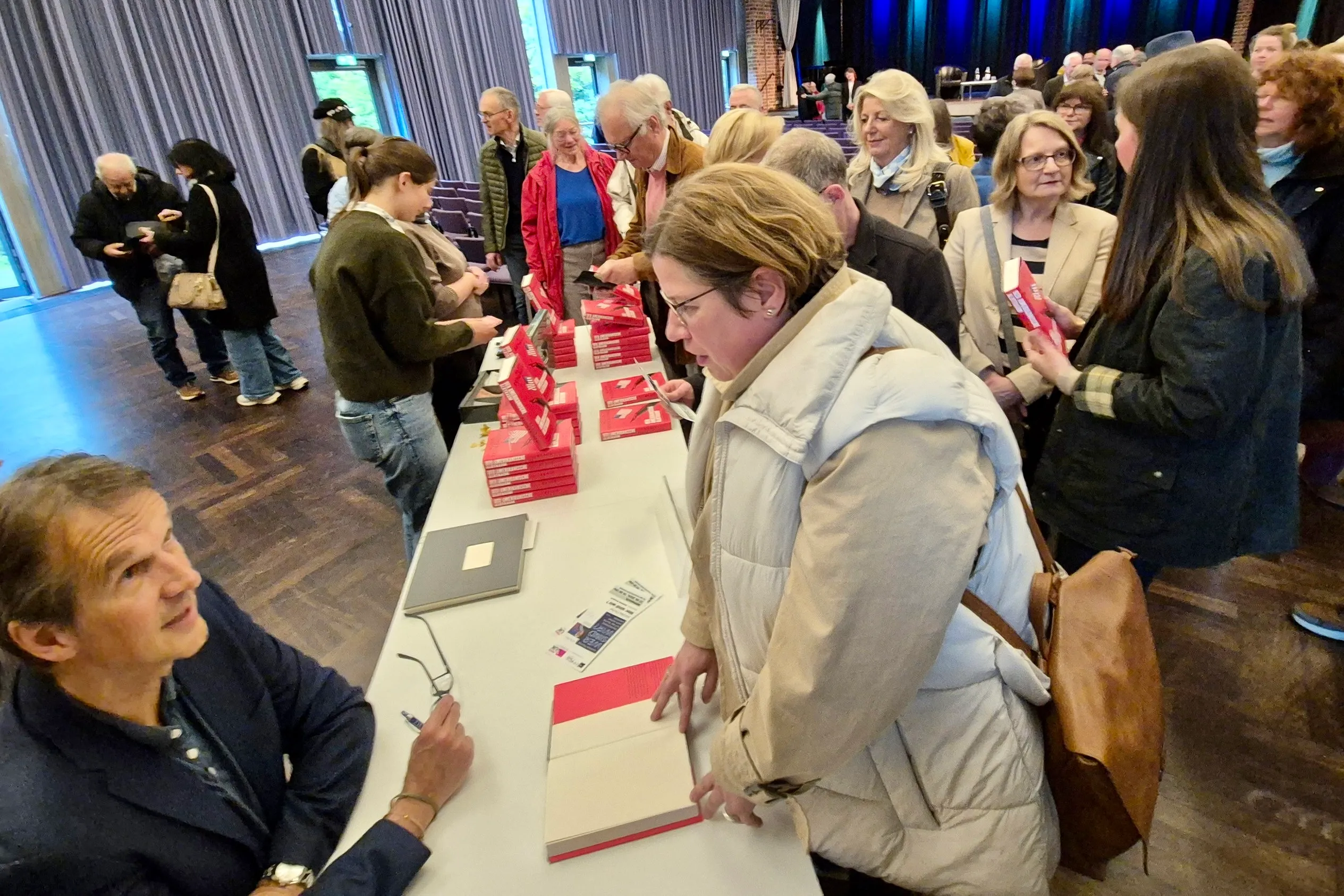 Klaus Brinkbäumer signiert Bücher an einem Tisch, umgeben von einer Gruppe von Menschen unterschiedlichen Alters.