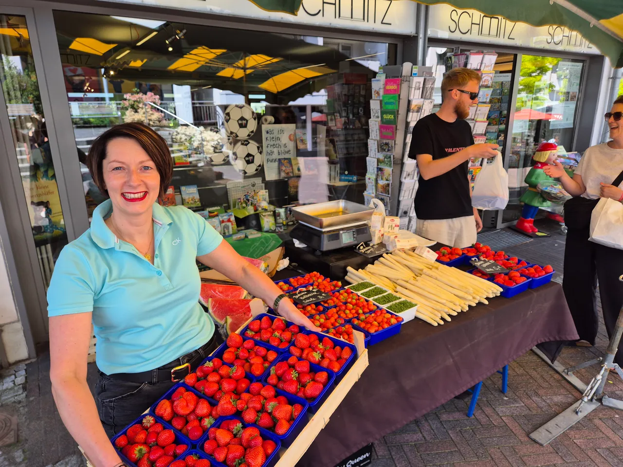Frau mit rotem Lippenstift steht lächelnd hinter einem Stand mit Erdbeeren und Spargel auf einem Markt in der Fußgängerzone. Im Hintergrund ein Mann mit Bart und weitere Marktbesucher.