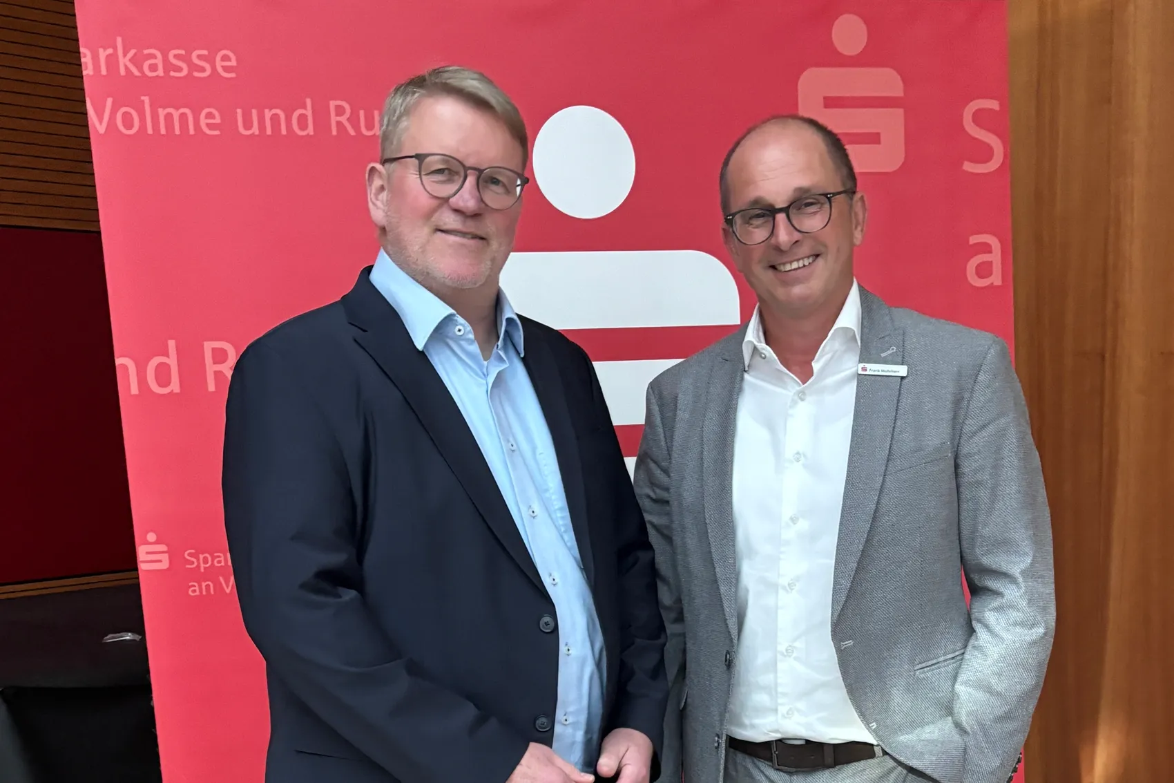 Zwei lächelnde Männer mit Brille im Anzug stehen vor rotem Hintergrund mit Sparkassen-Logo und Text. Links Jörg Klepper, rechts Frank Mohrherr.