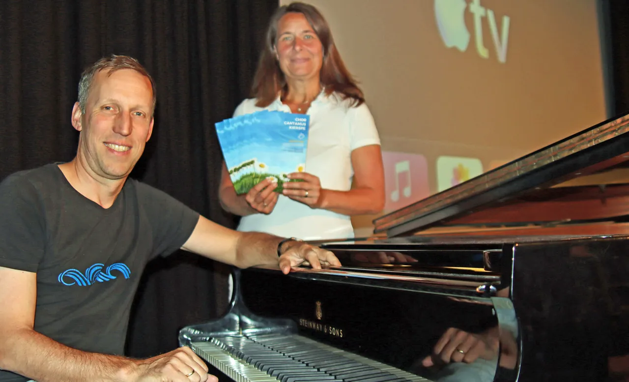 Chorleiter Michael Otto sitzt lächelnd vor einem Flügel, während Miriam Haarbach ein Programmheft für das Mittsommerkonzert des Chors Cantamus hält.