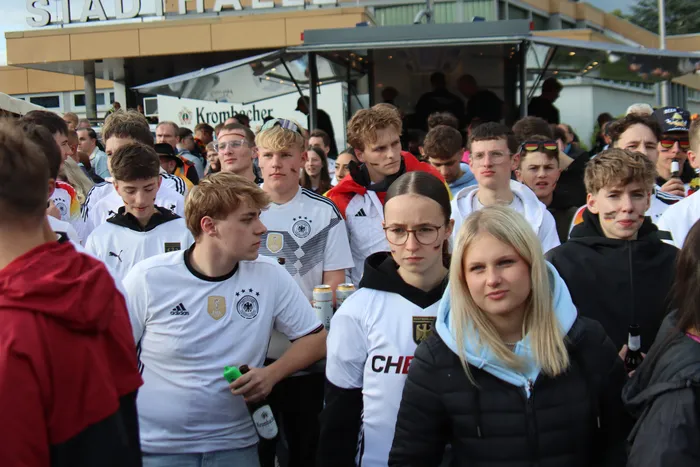Junge Menschen in deutschen Fußballtrikots und mit Deutschlandfahnen feiern ausgelassen auf dem Otto-Fuchs-Platz. Im Hintergrund ist ein Getränkestand zu sehen.