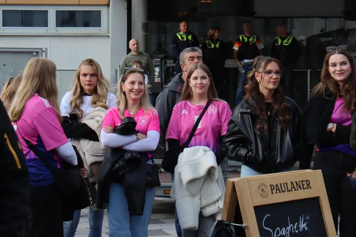 Eine Gruppe von Menschen, darunter viele junge Frauen in rosa Trikots, steht und lächelt in die Kamera. Im Hintergrund sind weitere Zuschauer und ein Schild mit der Aufschrift 