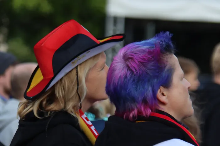 Zwei Fußballfans mit deutschen Farben im Haar und auf Hüten schauen aufgeregt zu. Im Hintergrund sind weitere Zuschauer bei einem Public Viewing zu sehen.