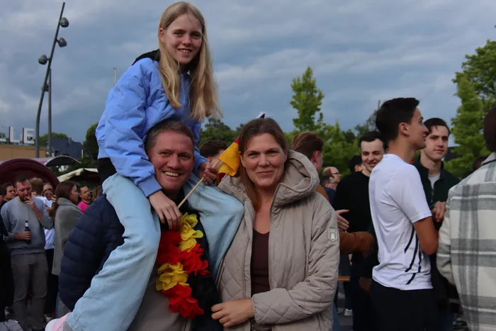 Familie feiert beim Public Viewing den Sieg der deutschen Nationalmannschaft: Ein Mann trägt seine Tochter auf den Schultern, während seine Frau lächelnd daneben steht. Beide tragen Deutschland-Fahnen und -Schmuck. Im Hintergrund jubeln weitere Zuschauer.