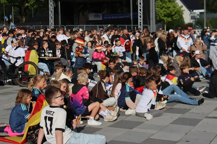 Eine große Menschenmenge sitzt und steht auf einem Platz und verfolgt ein Fußballspiel auf einer Leinwand. Viele Zuschauer tragen Trikots der deutschen Nationalmannschaft und schwenken Deutschlandflaggen.