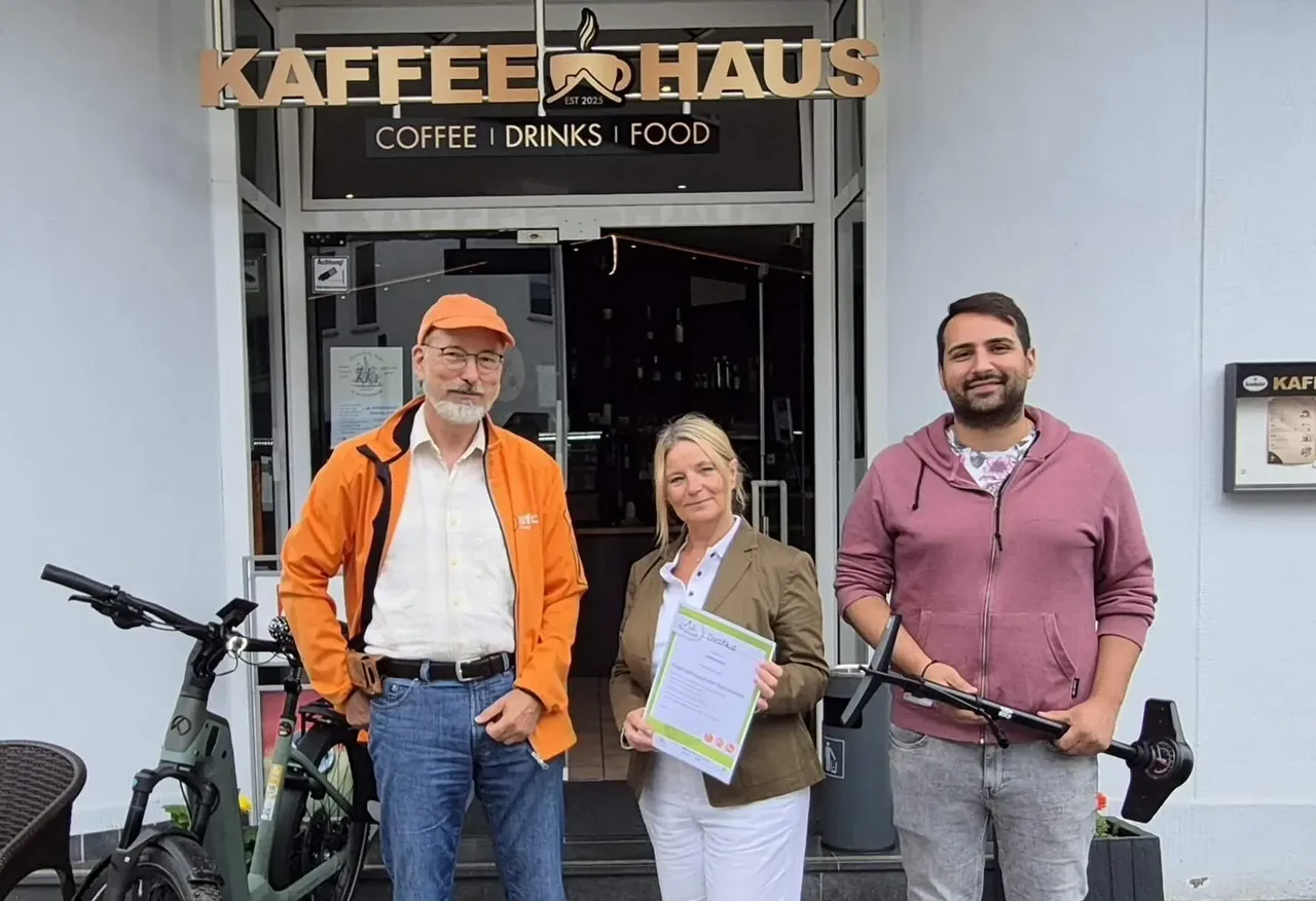 Sylvia Eick, Sebastian Rittner und Stefanos Triantafullou stehen vor dem Café Kaffeehaus. Sylvia Eick hält das Gütesiegel „Fahrradfreundliche Gastronomie“ in den Händen.