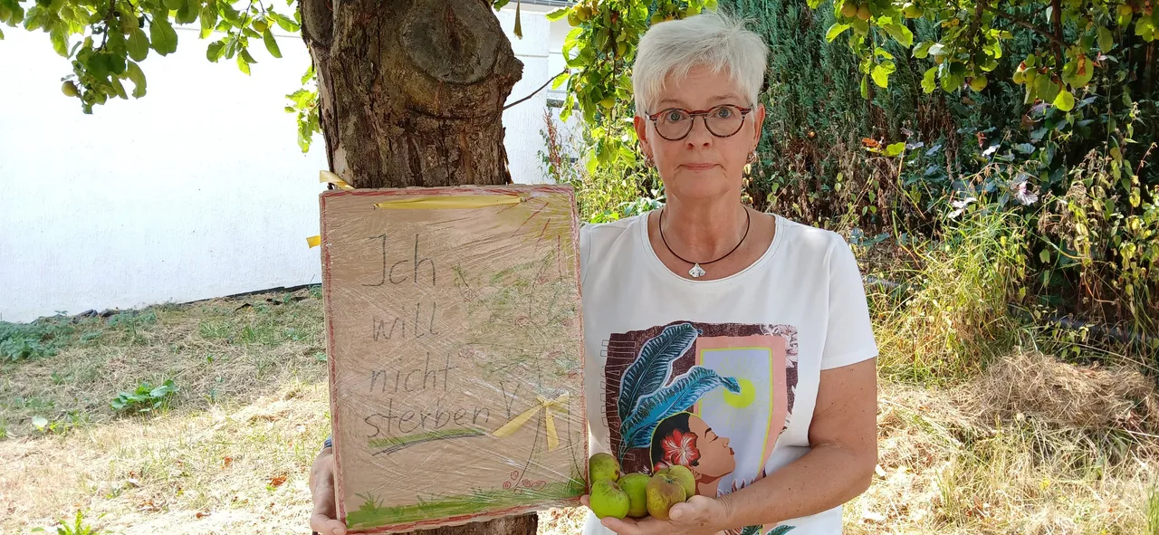 Eine Frau hält ein Holzschild mit der Aufschrift „Ich will nicht sterben“ und einige grüne Äpfel vor einem Baum.