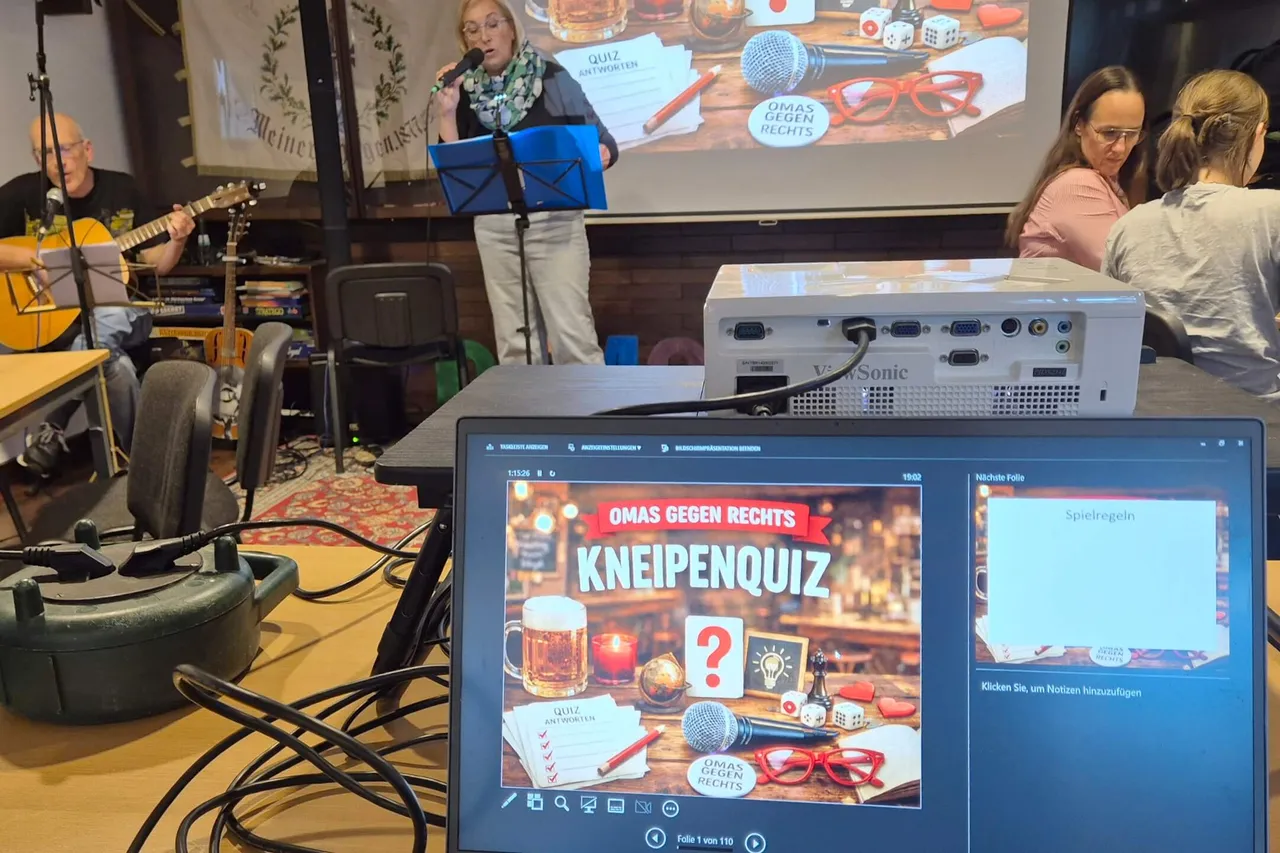 Eine Frau moderiert ein Kneipen-Quiz vor einer Leinwand mit dem Titel „OMAS GEGEN RECHTS KNEIPENQUIZ“, während im Hintergrund ein Musiker Gitarre spielt.