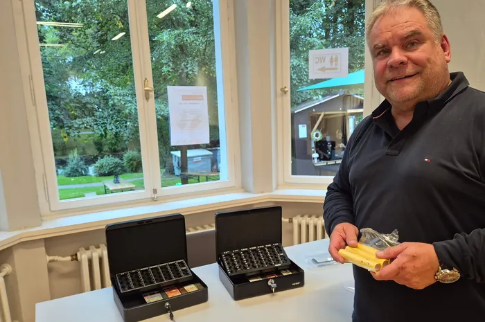 Ein lächelnder Mann in einem schwarzen Poloshirt hält ein kleines gelbes Paket vor zwei geöffneten schwarzen Etuis auf einem Tisch.