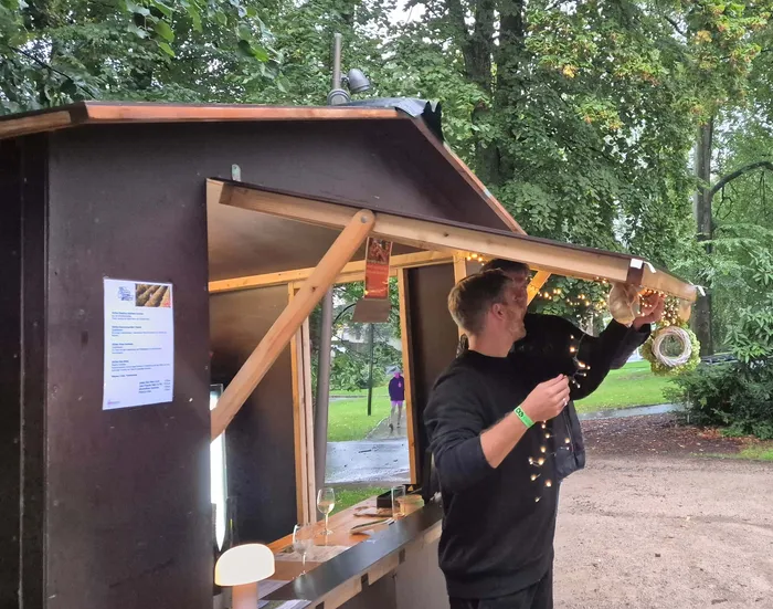 Ein Mann in einem schwarzen Sweatshirt steht an einem hölzernen Stand in einem Park, an dem Lichterketten hängen.