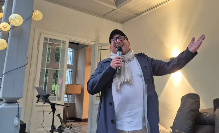 Ein lächelnder Mann mit Brille, Kappe und Schal spricht in ein Mikrofon und gestikuliert.