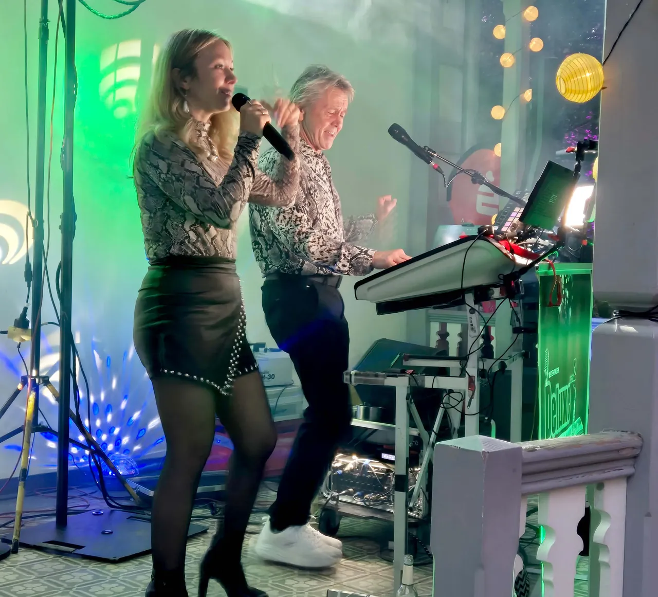 Eine Sängerin und ein Keyboarder treten bei einem Weinfest vor einer grün und blau beleuchteten Kulisse auf.