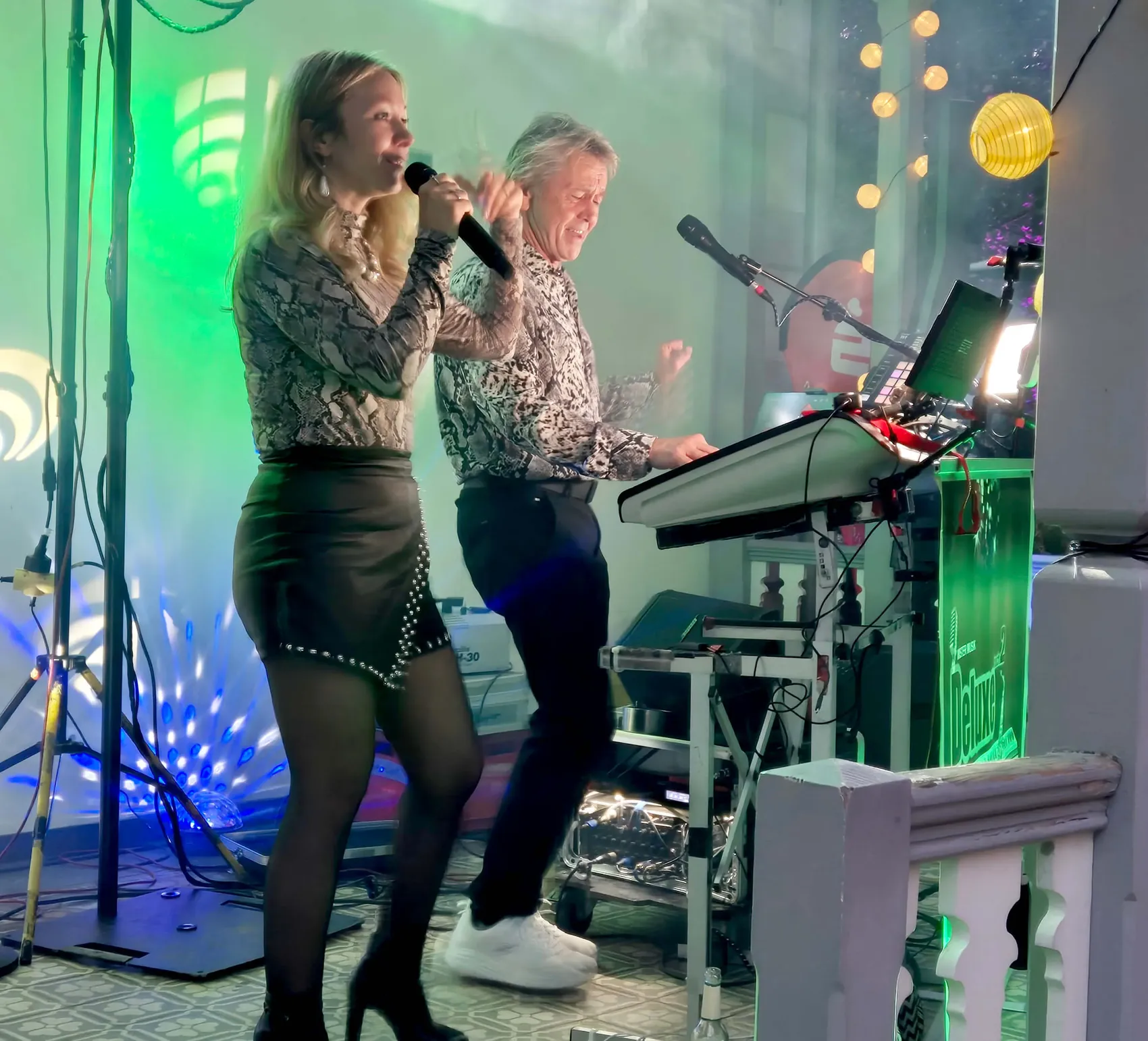 Eine Sängerin und ein Keyboarder treten bei einem Weinfest vor einer grün und blau beleuchteten Kulisse auf.