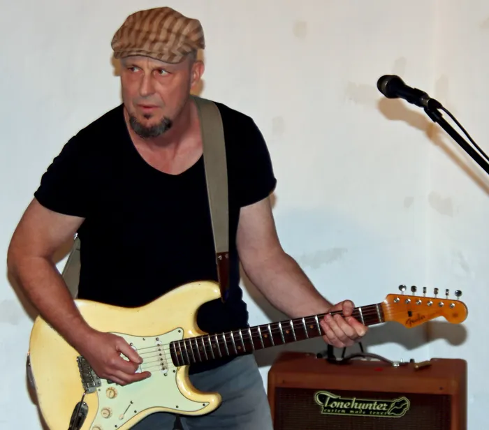 Chris Linder mit Schiebermütze spielt eine helle E-Gitarre vor einem Verstärker und einem Mikrofon.