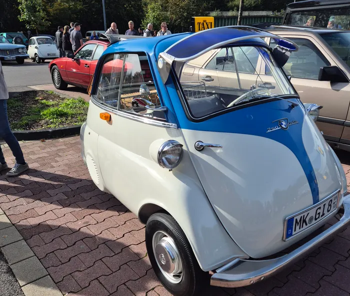 Ein weißer Kleinstwagen mit blauem Dach und blauem Frontstreifen steht auf einem gepflasterten Parkplatz bei einem Oldtimer-Treffen.