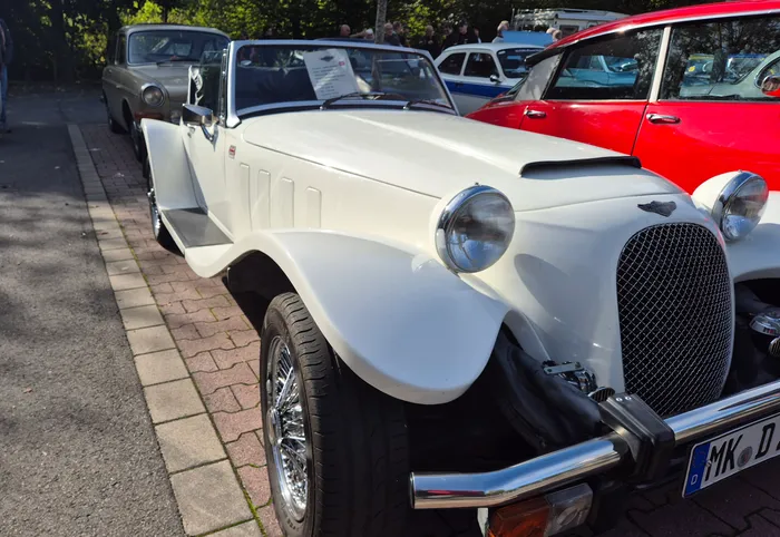 Ein weißes Oldtimer-Cabriolet mit runden Scheinwerfern und Chromdetails steht auf einem Parkplatz neben einem roten Auto.