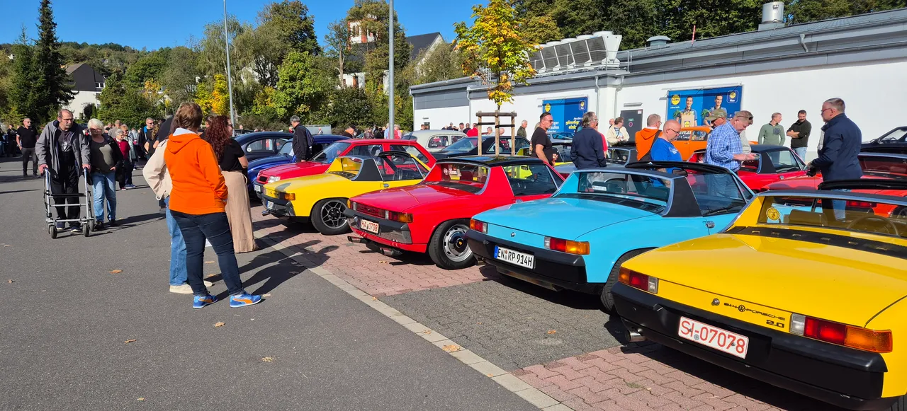 Eine Reihe bunter Oldtimer, darunter mehrere Porsche 914 in Gelb, Rot und Blau, stehen auf einem Parkplatz, während Menschen die Fahrzeuge betrachten.