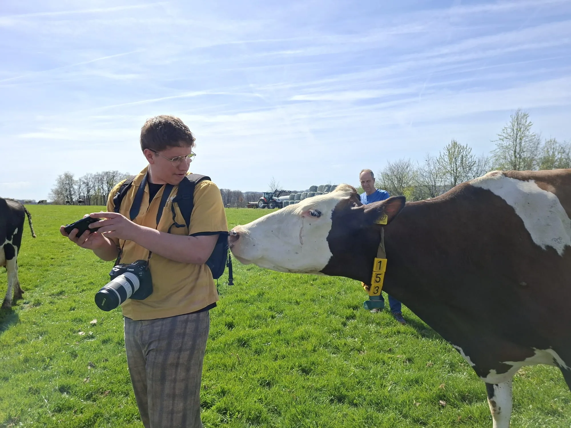 Ein junger Mann fotografiert eine schwarz-weiß gefleckte Kuh auf einer grünen Wiese. Im Hintergrund steht ein weiterer Mann und landwirtschaftliche Fahrzeuge sind zu sehen.