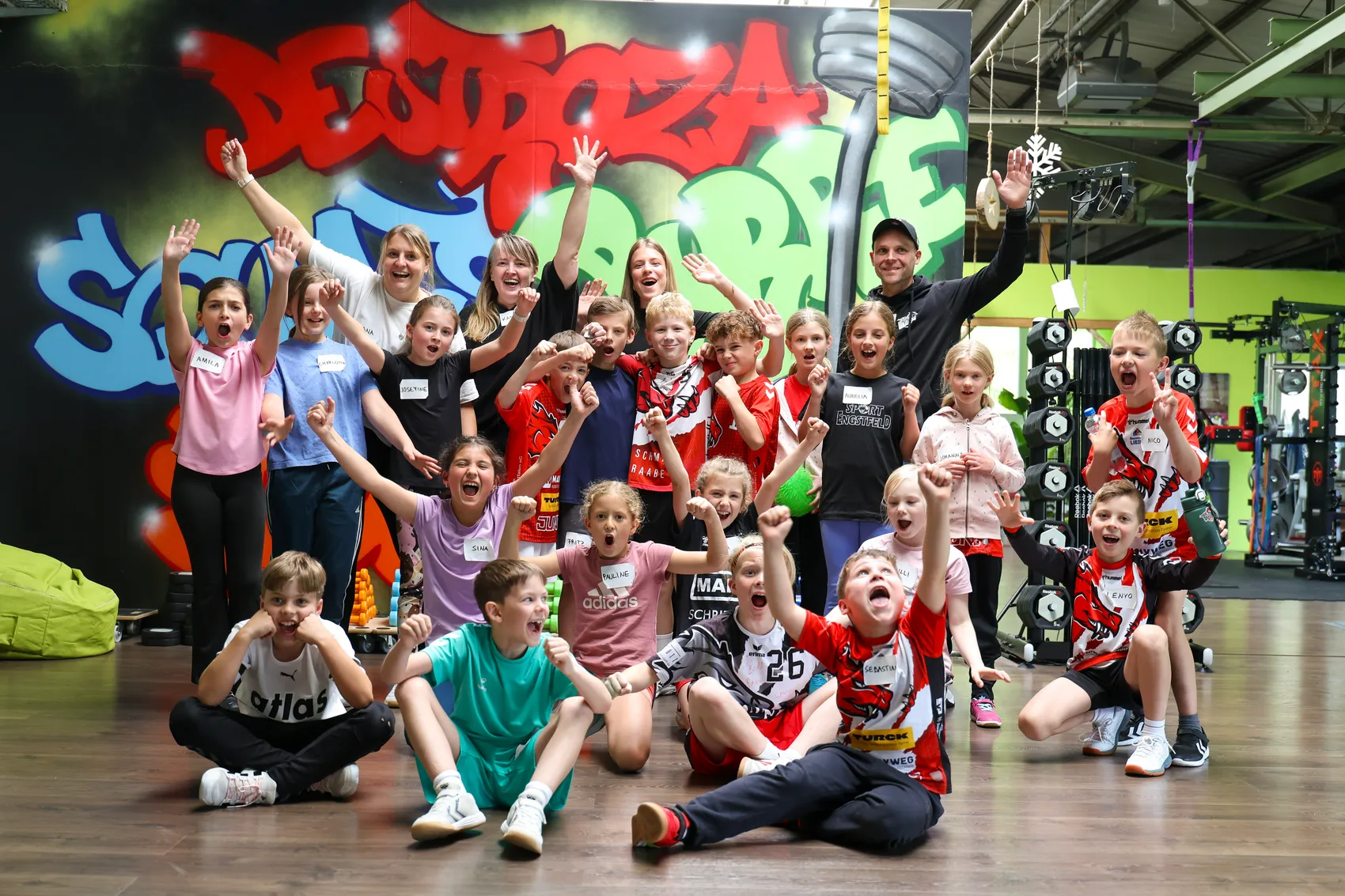 Gruppe von Kindern und Jugendlichen mit Betreuern im Fitnessstudio, die mit hoch erhobenen Armen jubeln. Viele tragen rote Trikots mit der Aufschrift „SGSH Dragons“. Im Hintergrund eine Wand mit Graffiti und Sportgeräte.