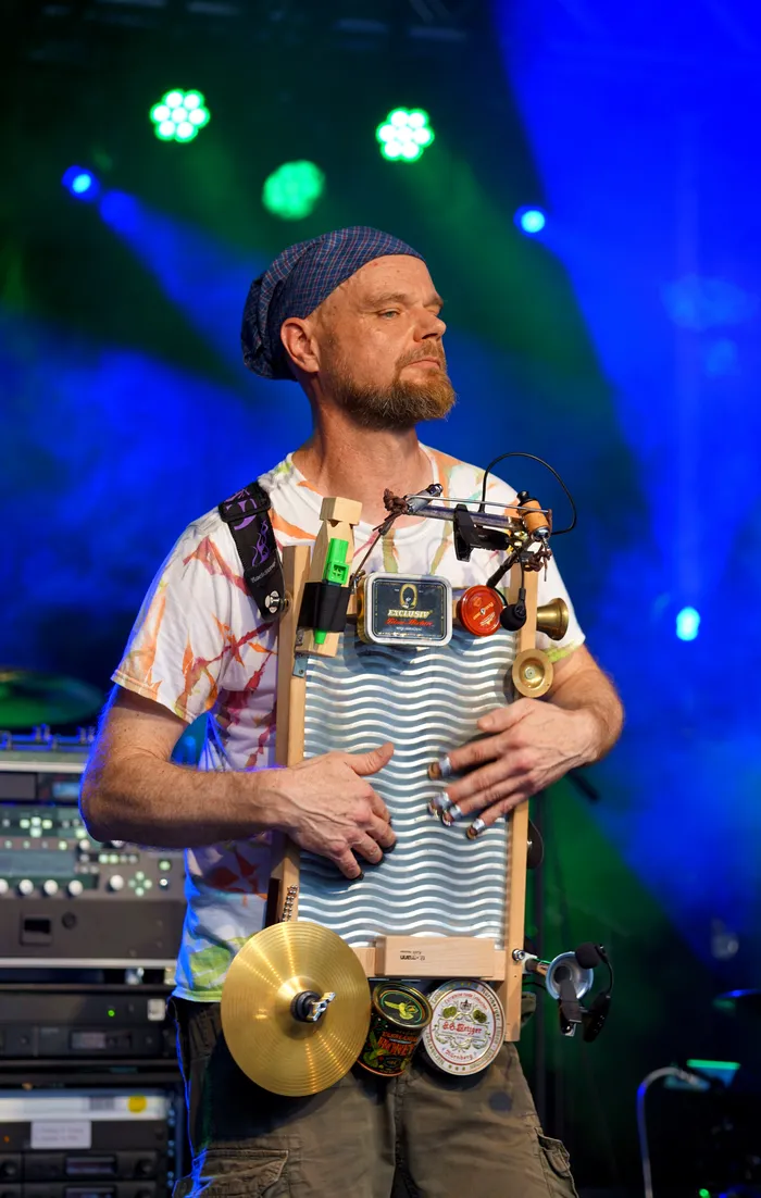 Ein Mann mit Bart und Mütze steht auf einer Bühne und spielt ein selbstgebautes Musikinstrument aus Holz und Metall. Das Instrument ist mit verschiedenen Gegenständen und kleinen Becken bestückt.