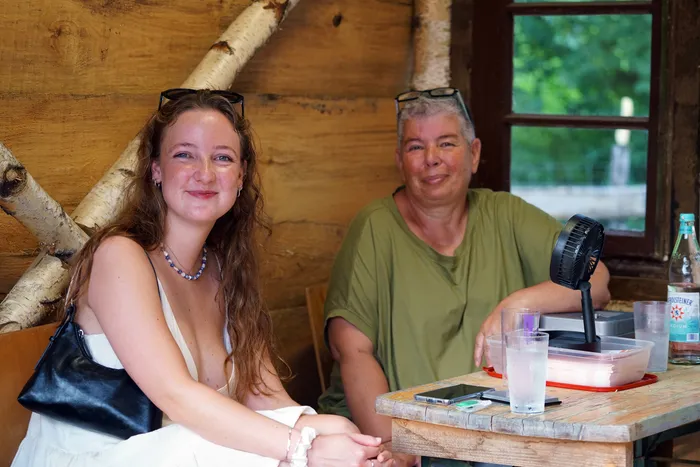 Zwei lächelnde Frauen sitzen an einem Holztisch. Die jüngere Frau links trägt eine Sonnenbrille auf dem Kopf und eine weiße Bluse, während die ältere Frau rechts ein grünes Poloshirt trägt. Ein kleiner Ventilator steht auf dem Tisch zwischen ihnen.