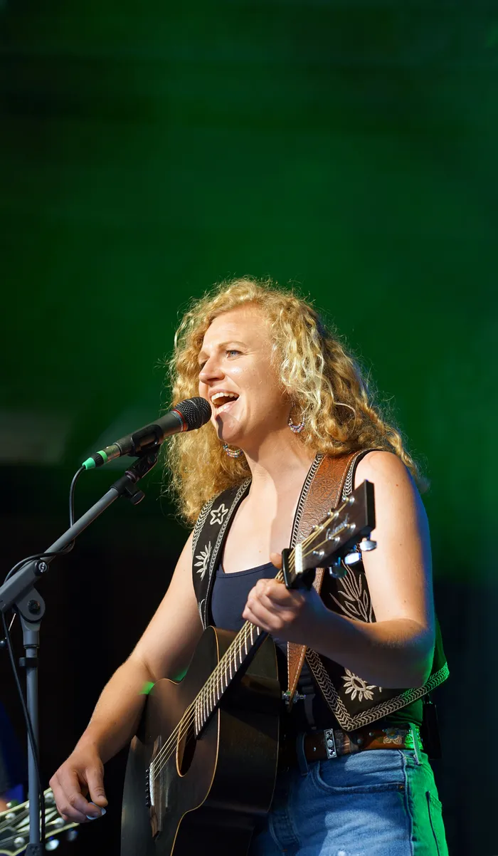 Eine Frau mit lockigem blonden Haar spielt Gitarre und singt in ein Mikrofon. Sie trägt ein schwarzes Top, Jeans und einen Ledergurt.