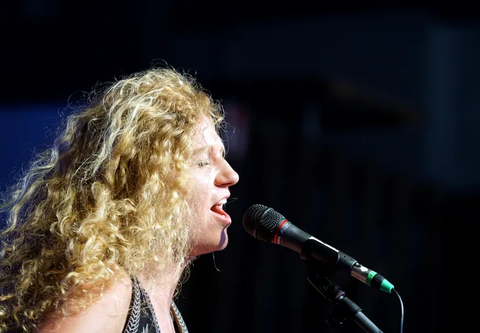 Eine Frau mit lockigem, blondem Haar singt in ein Mikrofon. Ihr Profil ist im hellen Licht zu sehen, der Hintergrund ist dunkel.