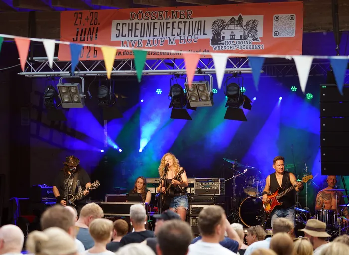Band spielt auf einer Bühne vor Publikum bei dem „Düsseldorfer Scheunenfestival“. Im Hintergrund ein Banner mit dem Festivalnamen und Datum. Blaue und grüne Bühnenbeleuchtung.