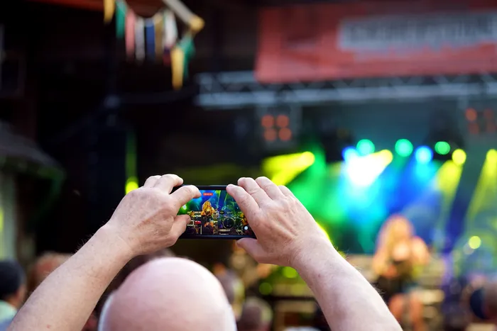 Eine Person filmt mit einem Smartphone eine Bühne bei einem Konzert oder einer Veranstaltung. Im Hintergrund sind Lichter und Menschen zu sehen.
