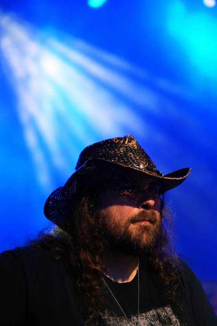 Ein Mann mit langem, lockigem Haar und Bart trägt einen dunklen Cowboyhut. Im Hintergrund ist blaues Licht zu sehen.