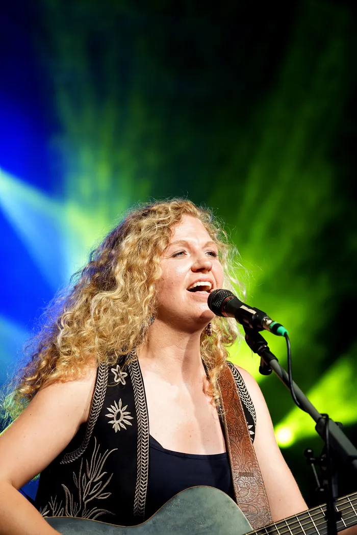 Eine Frau mit lockigem blondem Haar singt in ein Mikrofon, während sie eine Akustikgitarre spielt. Im Hintergrund ist eine bunte Bühnenbeleuchtung zu sehen.