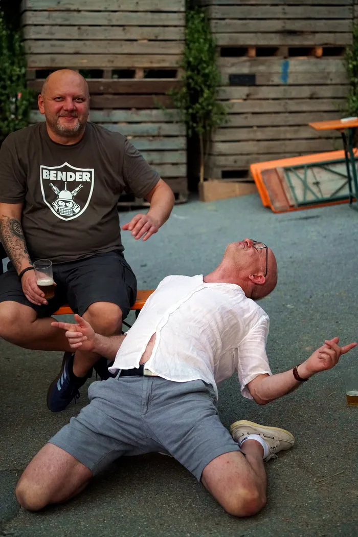 Ein Mann liegt auf dem Rücken, während ein anderer Mann ihm gegenüber sitzt und mit den Händen eine Geste macht. Im Hintergrund sind Holzelemente und Tische zu sehen.