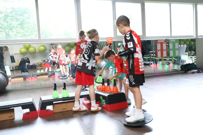 Kinder trainieren in einer Turnhalle. Vier Jungen stehen im Vordergrund und üben auf einer Balance-Plattform und kleinen Podesten, während im Hintergrund weitere Kinder und Erwachsene zu sehen sind.