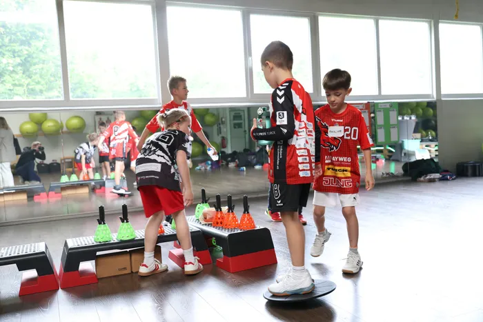 Junge Handballspieler trainieren Balance und Koordination in einer Turnhalle. Ein Junge steht auf einer Wippe, während ein anderer über eine niedrige Hürde steigt.