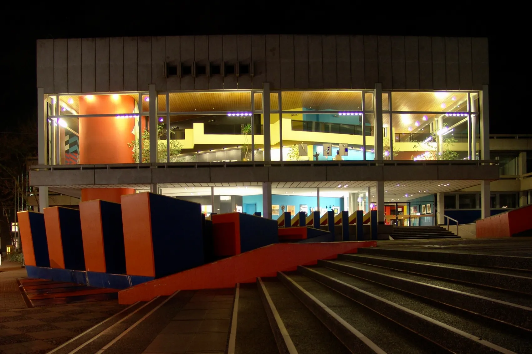 Das Schwelmer Kreishaus ist nachts teilweise beleuchtet, während ein Teil des Gebäudes im Dunkeln liegt. Vor dem Gebäude befinden sich farbige Skulpturen und eine Treppe.